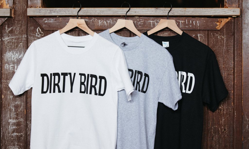 Three Dirty Bird t-shirts in white, gray and black hanging on the wall
