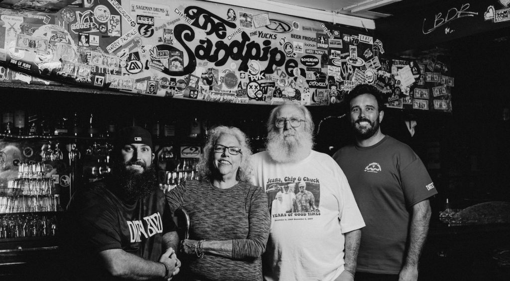 Four people standing together in front of the sticker-covered wall and the Sandpiper sign, black and white