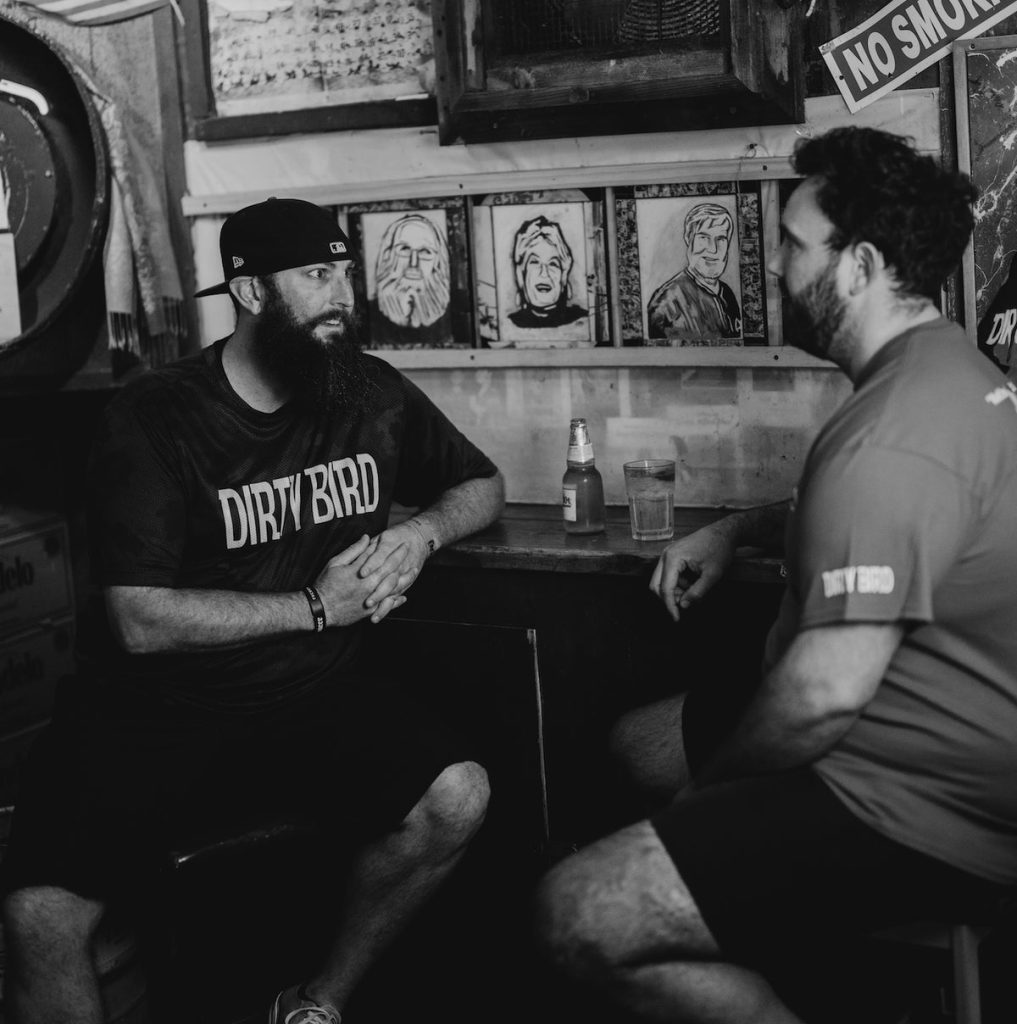 Two people in Dirty Bird shirts talking at a table under framed portraits, black and white